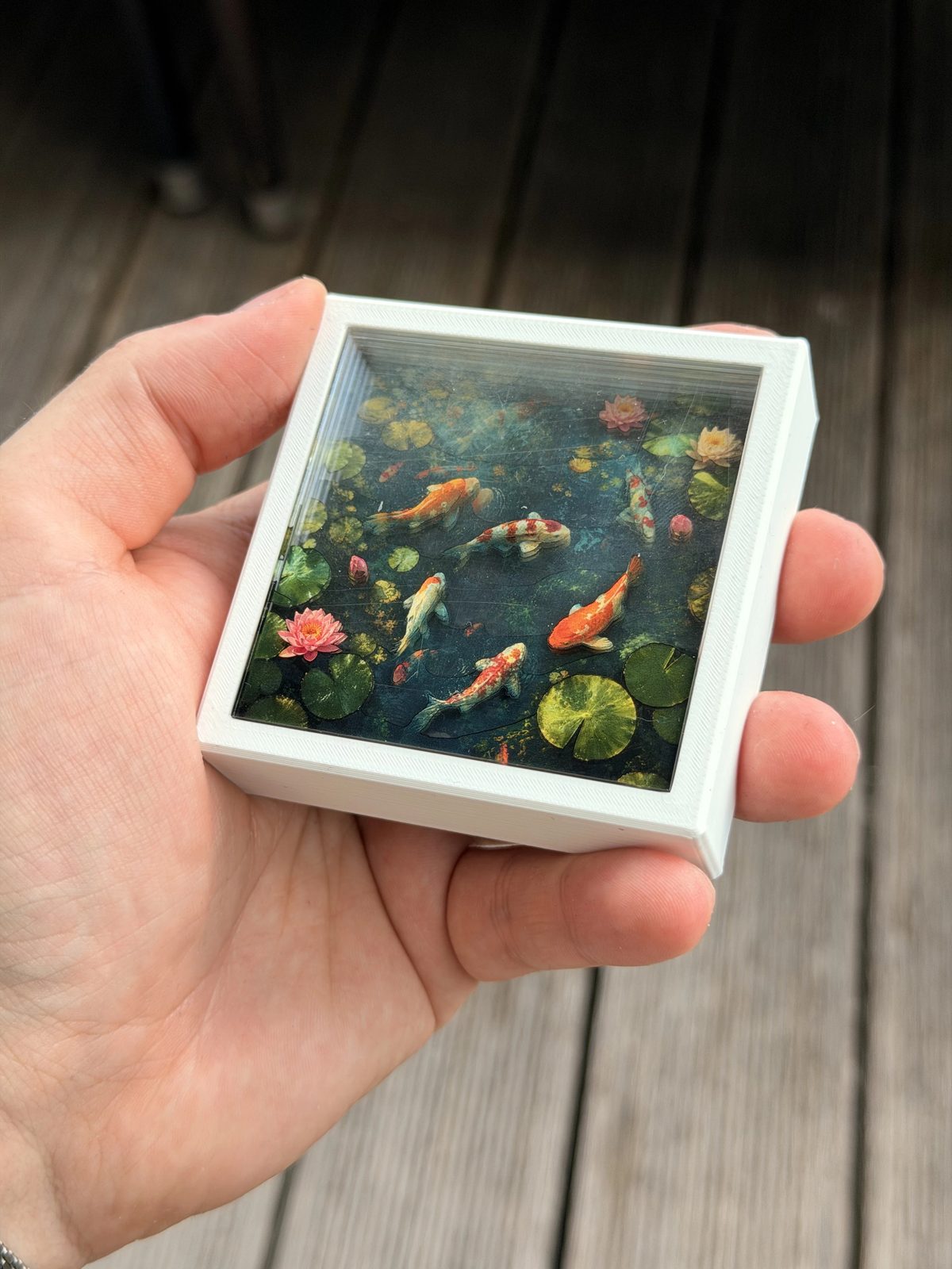 Hand holding a framed 3D koi pond artwork with layered water lilies.